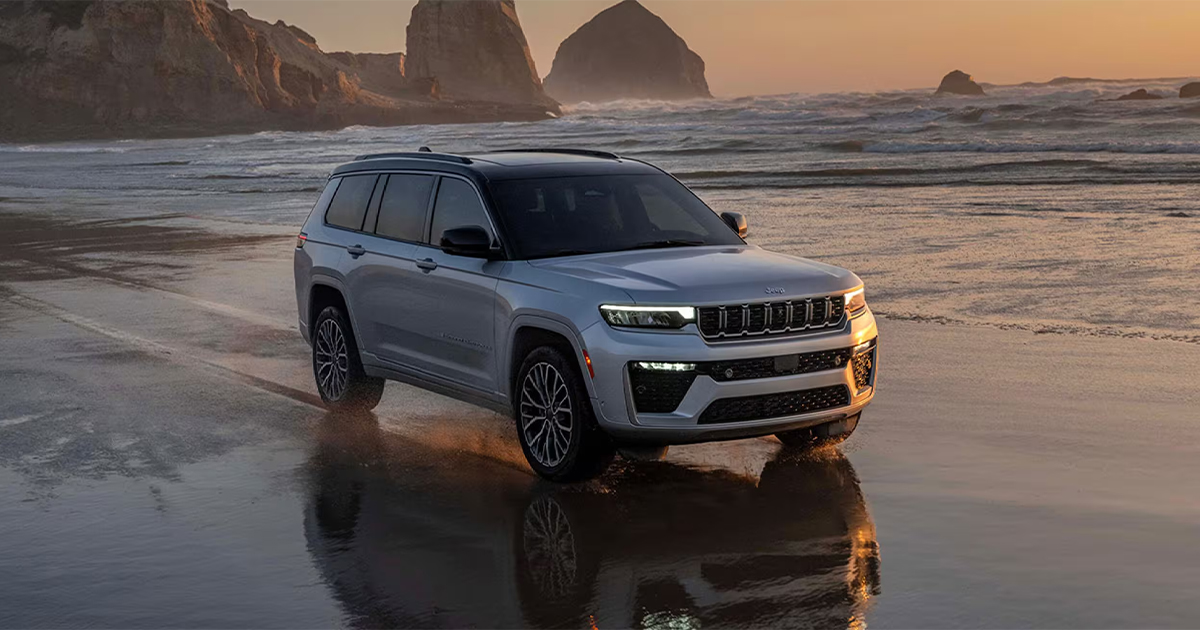 A silver 2026 Jeep Grand Cherokee L featuring a refreshed front fascia and multi-spoke alloy wheels on a wet coastal shore